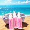 Three bottles labeled Shampoo, Conditioner, and Shower Gel with pink floral designs are placed on sandy beach next to a holographic pouch, ocean in background.