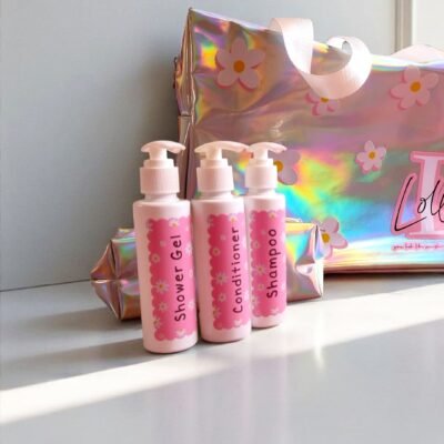Three pink bottles labeled shower gel, conditioner, and shampoo stand in front of a shiny holographic bag with floral designs, conveying a playful tone.