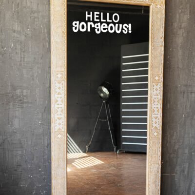 A tall, ornately framed mirror stands against a textured gray wall. The words "HELLO gorgeous!" are written on it. A spotlight and striped light patterns are reflected.