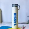 Tall white water bottle with "YASSIN" in bold blue letters, clear lid, and handle. Set on blue and yellow paper with scattered paper clips.