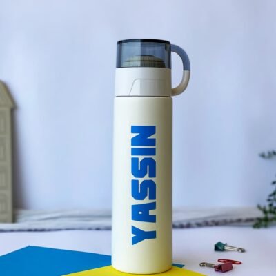 Tall white water bottle with "YASSIN" in bold blue letters, clear lid, and handle. Set on blue and yellow paper with scattered paper clips.