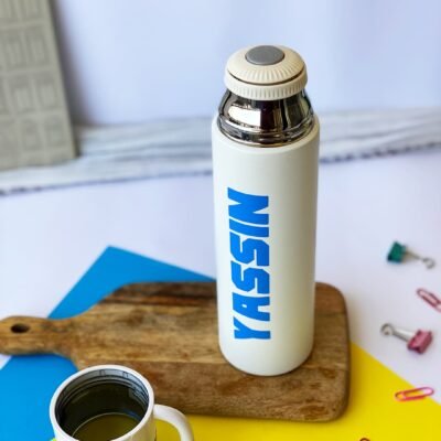 A white thermos with a blue name sits on a wooden board. Nearby, a black and white cup and colorful clips rest on a bright yellow and blue background.