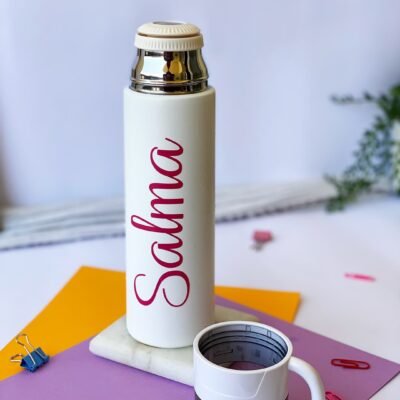 A white bottle with "Salma" in red script stands on a marble base. Beside it, a small mug rests on purple paper. Vibrant, creative workspace.