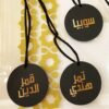 Three black circular tags with Arabic text and black cords lie on a surface. A decorative gold geometric pattern lies beneath, adding an elegant touch.