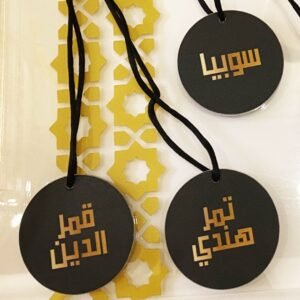 Three black circular tags with Arabic text and black cords lie on a surface. A decorative gold geometric pattern lies beneath, adding an elegant touch.