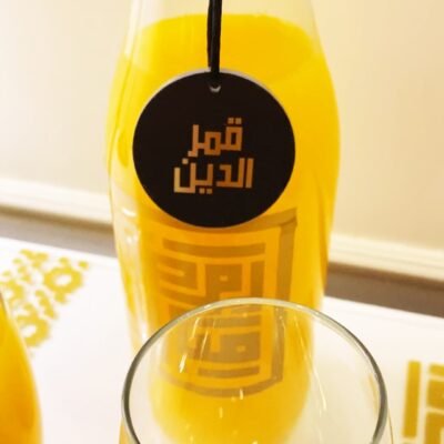 A clear bottle with orange apricot juice, labeled in Arabic, stands behind an empty glass. Bright, inviting color conveys a fresh, vibrant mood.
