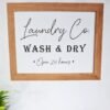 A sign with a wooden frame reads "Laundry Co. Wash & Dry Open 24 hours" in elegant black script, set against a white background. A plant decorates the lower left corner.
