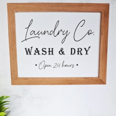 A sign with a wooden frame reads "Laundry Co. Wash & Dry Open 24 hours" in elegant black script, set against a white background. A plant decorates the lower left corner.