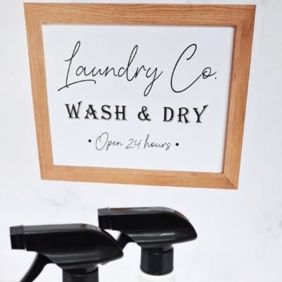 Wood-framed sign reads "Laundry Co. Wash & Dry Open 24 hours" on a white wall. Two white spray bottles with black nozzles sit in the foreground.