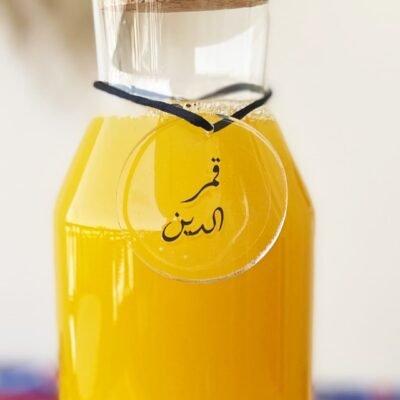 A clear glass bottle with a cork stopper contains bright yellow liquid. A round tag with Arabic text hangs from the neck, creating a fresh, inviting feel.