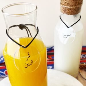 Two glass bottles on a white surface. The front bottle contains bright orange juice, and the back has a white liquid with a cork stopper. Labels with Arabic calligraphy hang from each. A colorful fabric with red and blue patterns is partially visible in the background.