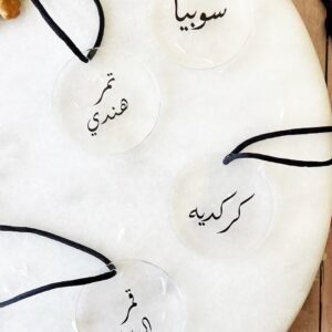 Four round, transparent tags with black Arabic calligraphy are displayed on a white surface. Each tag has a black cord attached, creating an elegant and artistic presentation.