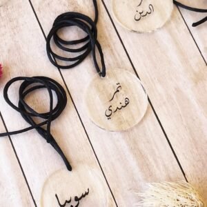 Clear round pendants with Arabic calligraphy lie on a light wooden surface. Black cords are attached to each, adding elegance and simplicity.