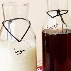 Two glass bottles with round labels in Arabic script. The left bottle contains sobia, while the right holds carob juice. Simple and elegant.