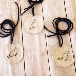 Three transparent round tags with black cords rest on a wooden surface. Each tag features Arabic calligraphy. The scene is calm and minimalist.