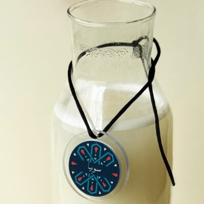 Glass bottle filled with a creamy beverage, adorned with a decorative circular tag featuring geometric patterns and Arabic script, evoking a serene and artistic vibe.