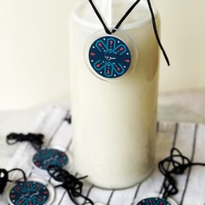 Clear glass bottle filled with a creamy white drink, featuring a decorative blue and red circular tag. Several similar tags lie nearby on a striped cloth.