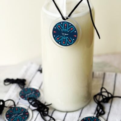 Clear glass bottle filled with a creamy white drink, featuring a decorative blue and red circular tag. Several similar tags lie nearby on a striped cloth.