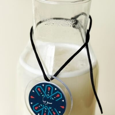 A clear glass jug filled with a creamy white liquid is adorned with a round pendant. The pendant features a geometric blue and red design, hanging by a black string.