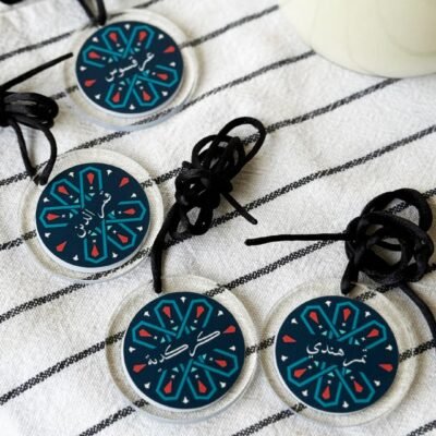 Four round keychains with colorful geometric patterns and Arabic script are on a striped cloth. They have black cords, creating an artisanal feel.
