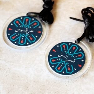 Two circular danglers with geometric patterns and Arabic script, featuring teal, red, and white on a dark blue background, lie on a marble surface.