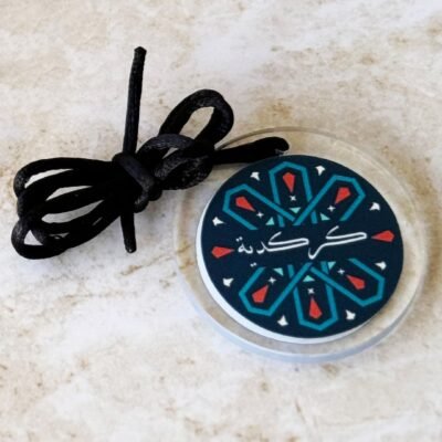 Round pendant with colorful geometric patterns and Arabic script on a stone surface. Black cord tied alongside, creating a traditional, artistic feel.
