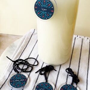 A tall glass bottle of milk with a blue, patterned tag hangs around its neck on a striped cloth. Three similar tags with black strings lie nearby.