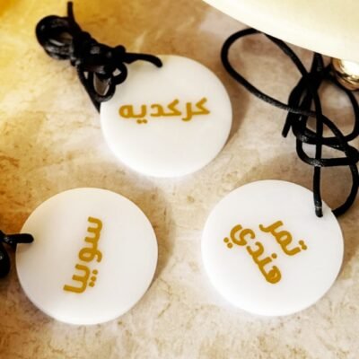 Three round white pendants with Arabic script in gold are tied with black cords on a beige marble surface. The scene conveys elegance and cultural richness.