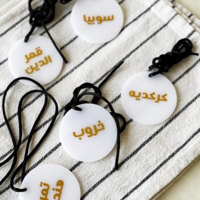 Five round white tags with Arabic text in gold, each attached to a black cord, are arranged on a striped fabric, creating an organized and elegant display.