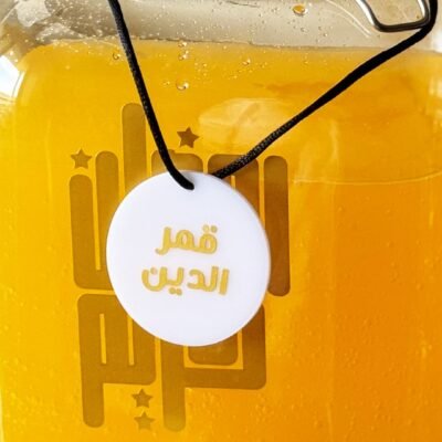 A clear jar filled with vibrant orange liquid, sealed with a latch. A round white tag with Arabic script hangs from a black cord around the jar’s neck.