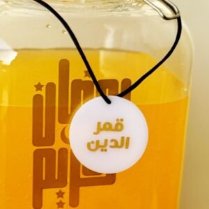 A glass jar filled with a bright orange liquid, sealed with a metal clasp. Arabic text is printed on the jar and a round tag hangs on a string.