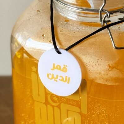 Close-up of a glass jar filled with orange liquid, labeled in Arabic. A white tag hangs by a black cord. The scene suggests a traditional beverage.
