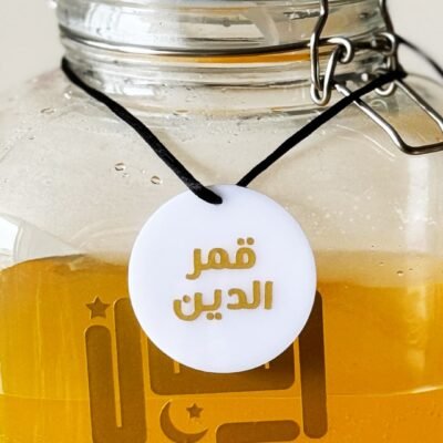 A glass jar filled with golden orange liquid, featuring Arabic calligraphy and a round white tag with Arabic text. The jar has a metal clasp.