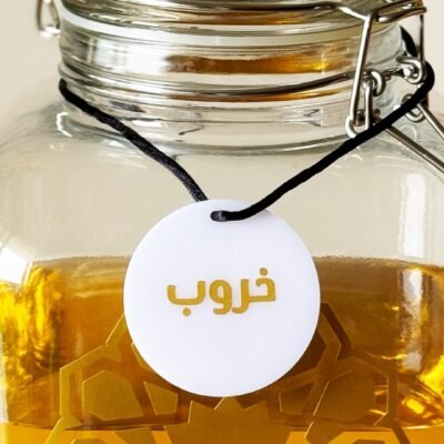 Glass jar with a metal clasp, filled with golden liquid. A white tag with Arabic writing and black string hangs around the jar, suggesting a homemade product.