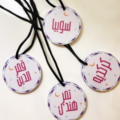 Four round white tags with colorful borders and Arabic text hang from black strings. Each tag features small crescent and star decorations.