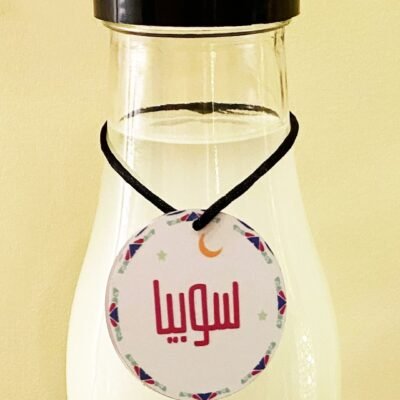 A clear glass bottle with a black cap contains a white liquid. A round tag with Arabic text and decorative patterns hangs around its neck.