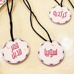 Three round badges with Arabic text hang on black cords. Each badge features a colorful border, crescent moon, and is placed on a beige surface.