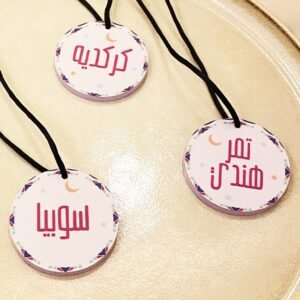 Three round tags with Arabic text hang on black cords. Each tag features a decorative border with small stars and a crescent moon, placed on a smooth, beige surface.