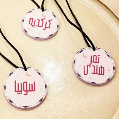 Three round tags with Arabic text hang on black cords. Each tag features a decorative border with small stars and a crescent moon, placed on a smooth, beige surface.