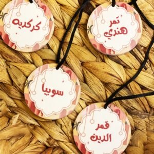 Round tags with Arabic calligraphy hang on black cords against a woven straw mat. Each tag features decorative red patterns, creating a warm, cultural vibe.