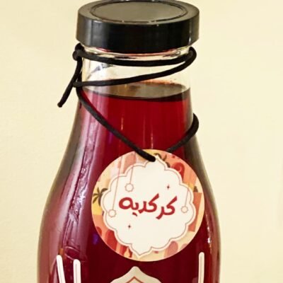 A glass bottle filled with dark red liquid features a black screw cap and a decorative label tied with string, displaying Arabic script in white.