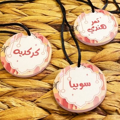 Three round tags with Arabic text hang on black strings over a woven straw surface. The design includes decorative pink and white borders.