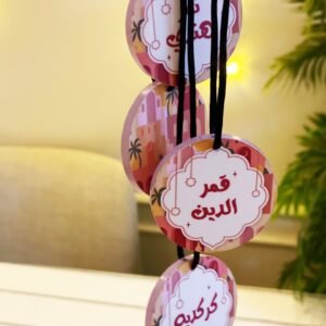 Four round, colorful tags with Arabic calligraphy hang from black strings. Background features soft lighting and green foliage, creating a warm, festive ambiance.