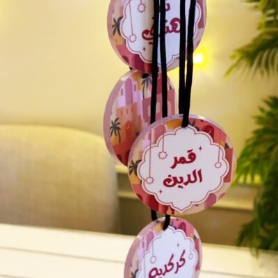 Four round, colorful tags with Arabic calligraphy hang from black strings. Background features soft lighting and green foliage, creating a warm, festive ambiance.