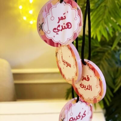 Round name tags with Arabic text hang on black strings against a backdrop of blurred green foliage and soft yellow fairy lights, creating a festive atmosphere.
