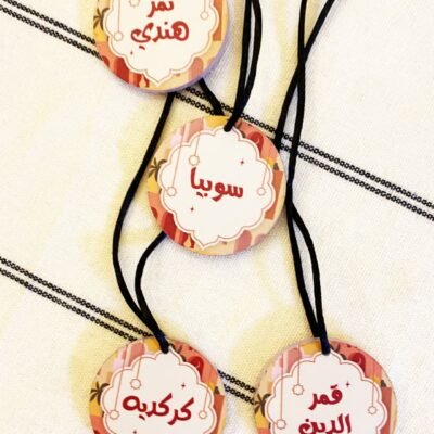 Four round tags with Arabic text hang from black strings on a white fabric background with black stripes. The design is ornate, colorful, and festive.