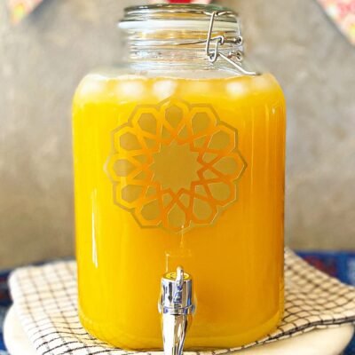 A glass drink dispenser filled with iced mango juice sits on a checkered cloth. A decorative pattern and festive bunting create a cheerful atmosphere.