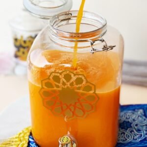 A glass jar with a spout is being filled with orange liquid, decorated with a geometric pattern. It rests on a blue and yellow ornate cloth.
