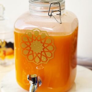 A clear glass jar filled with bright orange juice sits on a white counter. It features a metal spigot and a decorative geometric design, exuding a refreshing, inviting tone.