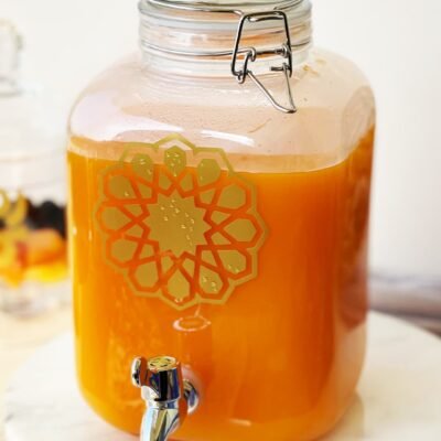 A clear glass jar filled with bright orange juice sits on a white counter. It features a metal spigot and a decorative geometric design, exuding a refreshing, inviting tone.